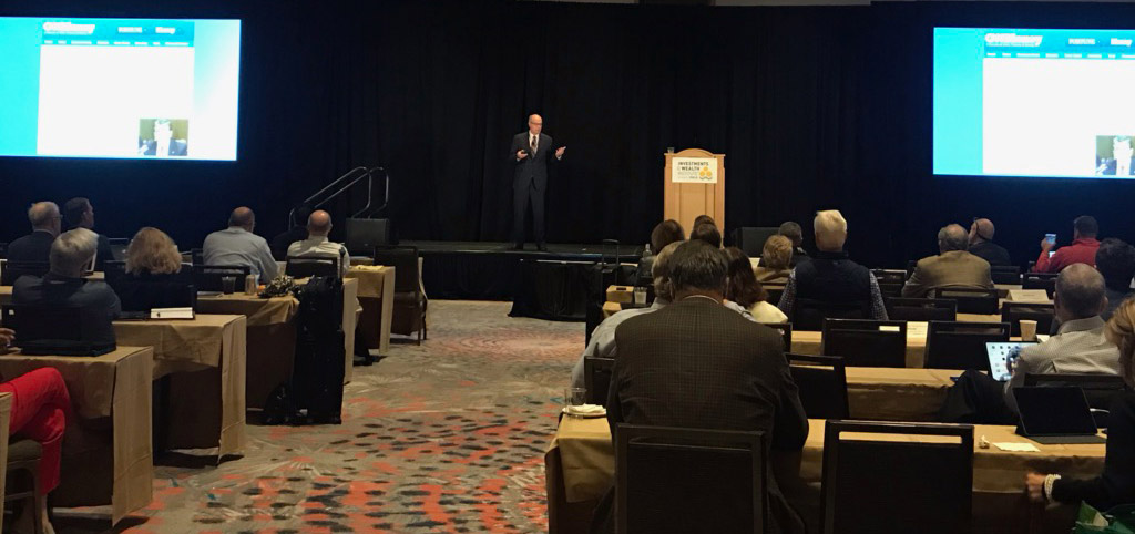 Image of Richard M. Bowen speaking on a stage before a crowd seated at tables.