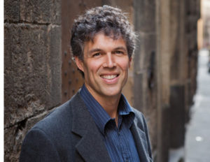 Image of Tom Mueller in gray suite standing against large bricks.