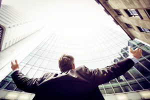 Low angle image of a man with his arms outstretched looking up at a tall glass buildling.